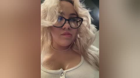 Media: Video of a curvy, fair-skinned woman with blonde hair and black-rimmed glasses, wearing a white ribbed tank top, standing indoors.