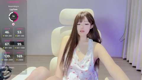 Media: Video of a young Asian woman with long black hair, wearing a white floral nightgown, sitting on a white chair, in a minimalist room.