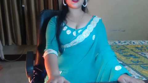 Media: Video of a South Asian woman in a teal sari with floral embroidery, seated in a black chair, wearing large hoop earrings, in a dimly lit room with beige walls and a bed with yellow and blue bedding.