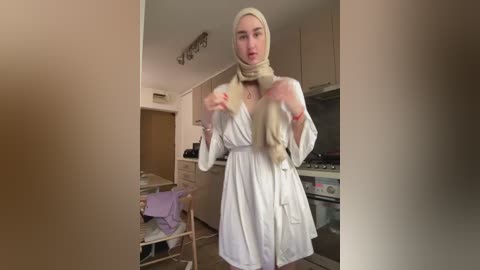 Media: Video of a woman in a white robe with a beige scarf, holding a long blonde wig, in a modern kitchen with beige cabinets and stainless steel appliances.