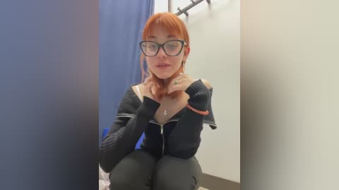 Media: Video of a pale-skinned, red-haired woman with glasses, wearing a black knitted sweater, sitting in a changing room. She touches her throat, looking thoughtful. Background shows a white wall and a blue curtain.