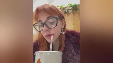 Media: Video of a fair-skinned woman with red hair, wearing black glasses and a purple fur coat, drinking from a white cup with an orange straw, in a blurred indoor setting.