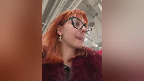 Media: Video of a young woman with shoulder-length, vibrant red hair, wearing black-framed glasses and a maroon fur coat, set against a blurred indoor background with white and beige tones.