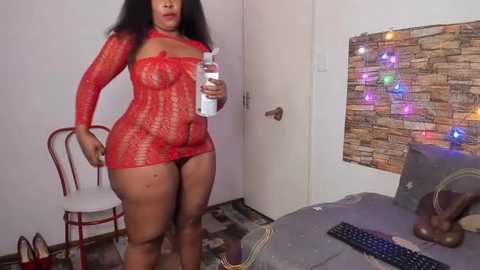 Media: Video of a curvy Black woman in red lace lingerie, holding a water bottle, standing in a modestly decorated room with a desk and chair.