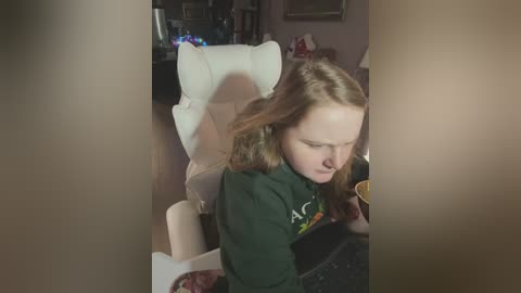 Media: Video of a young girl with shoulder-length brown hair, wearing a green shirt, sitting in a white recliner, eating a bowl of food, in a dimly lit living room with a TV and Christmas lights in the background.