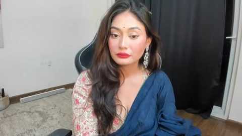 Media: Video of a South Asian woman with long, wavy dark hair, fair skin, and striking makeup, wearing a traditional saree with intricate patterns. She sits on a wooden floor, against a plain white wall and black curtain.