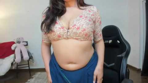 Media: Video of a curvy woman with medium skin tone, wearing a floral bra and blue skirt, in a simple room with a black gaming chair and stuffed animals.