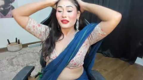 Media: Video of a South Asian woman with fair skin, long dark hair, and red lipstick, wearing a blue sari and floral top, sitting in a black office chair, with a beige rug and black curtain in the background.