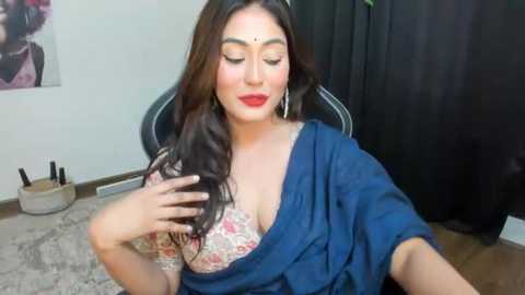 Media: Video of a South Asian woman with long dark hair, wearing a blue saree, floral bra, and red lipstick, sitting on a chair in a room with a black curtain, makeup brushes, and artwork on the wall.