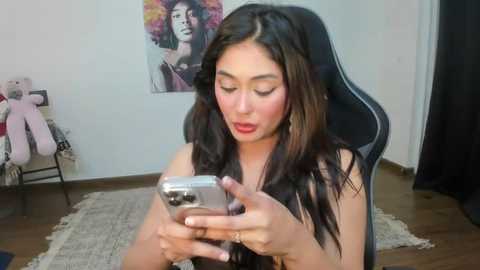 Media: Video of a young woman with long, dark hair, seated in a black gaming chair, wearing a sleeveless top, holding a silver phone, surrounded by stuffed animals and colorful wall art in a cozy room.