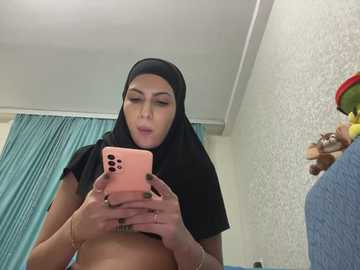 Media: A video shows a young woman with light skin, wearing a black hijab, topless, using a pink smartphone to take a selfie. Background includes a teal curtain and a textured wallpaper.