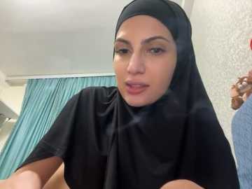 Media: Video of a light-skinned woman with a headscarf, wearing a black robe, standing in a bathroom with teal curtains and a floral-patterned wall.