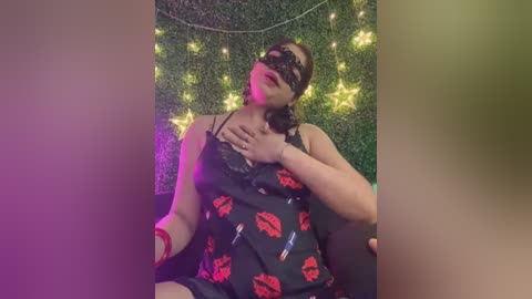 Media: Video of a plus-sized woman with light skin, wearing a black mask, red lipstick, and a black nightgown adorned with red lipstick prints. She sits against a green wall with fairy lights.