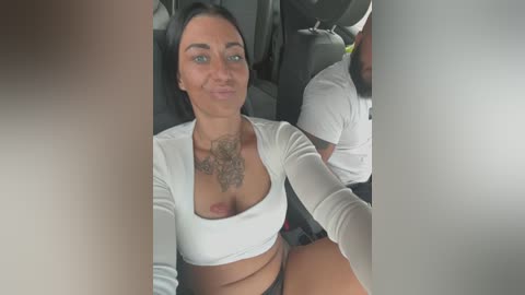 Media: Video of a woman with dark hair, medium skin tone, and tattoos on her chest, wearing a white crop top and black panties, seated in a car.