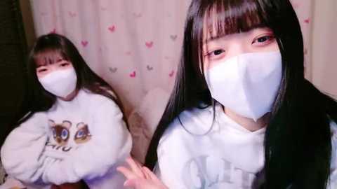 Media: Video of two Asian women in white face masks, one with straight black hair and the other with bangs, wearing white shirts with cartoon character prints. Background features pink heart-patterned curtains.