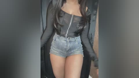 Media: Video of a woman with long, dark hair wearing a black, sheer long-sleeve top and high-waisted, light-wash denim shorts, standing in a dimly lit room with dark walls.