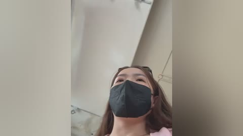 Media: Video of a young Asian woman with long brown hair, wearing a black face mask and pink jacket, looking up from a low angle. Background features beige walls and a bathroom sink.