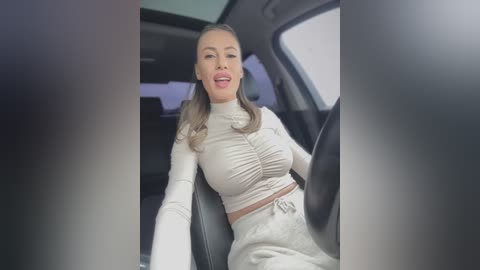 Video of a light-skinned, blonde woman in a white, ruched crop top and matching pants, smiling in a car seat with a window view.