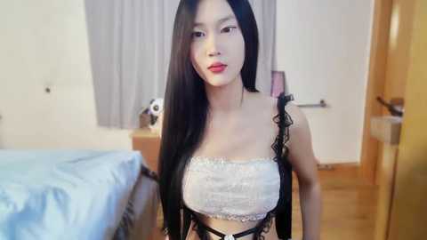 Media: Video of an Asian woman with long black hair, wearing a white lace bralette and black lace trim top, standing in a simple bedroom with a bed and wooden floors.