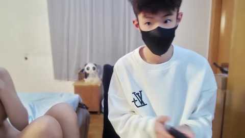 Media: Video of a young Asian man with short black hair, wearing a white sweatshirt, black face mask, and black shorts, playing a video game in a modern bedroom with wooden furniture, light grey curtains, and a white cat on a bed.