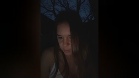 Media: A dimly lit video of a young woman with long brown hair, wearing a white tank top, looking directly at the camera with a serious expression. Bare trees are visible in the background against a dark sky.