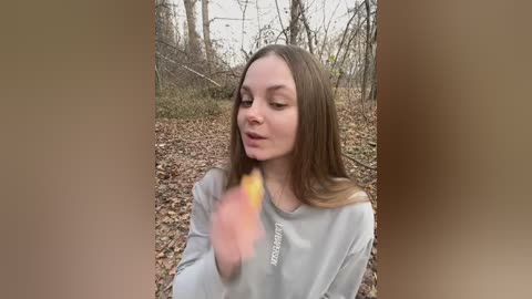 Media: Video of a young Caucasian woman with long brown hair, wearing a gray sweater, holding a glowing yellow stick in a forest with bare trees and brown leaves.