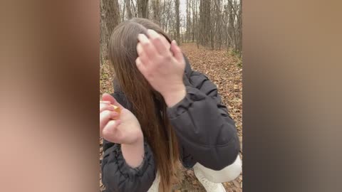 Media: A video captures a person with long brown hair kneeling on a forest floor, covering their face with both hands, wearing a black jacket and white pants.