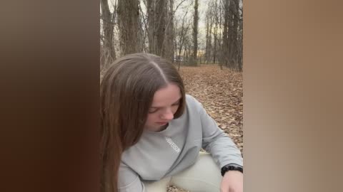 Media: Video of a young woman with long, straight brown hair, wearing a light gray sweatshirt, crouching on a bed of brown leaves in a forest. The background features leafless trees and a dirt path.