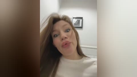 Media: A video captures a Caucasian woman with straight, light brown hair, making a pouty face, in a white top. She stands in a hallway with white walls, a framed black-and-white photo, and a door.