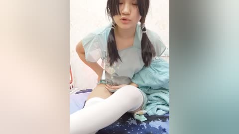 Media: Video of an Asian woman in a blue hospital gown, with pigtails, sitting on a bed with a blue blanket, looking down and touching her belly, in a dimly lit room.