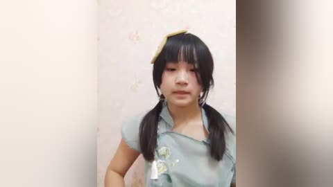 Media: A video of an Asian woman with straight black hair in pigtails, wearing a green dress with floral embroidery. She's standing in a light-colored room with a floral wallpaper, holding a yellow object above her head.