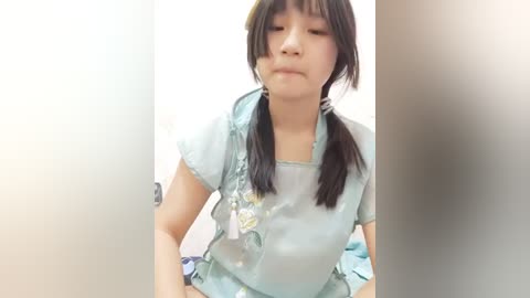 Media: Video of a young East Asian woman with straight black hair in pigtails, wearing a light blue floral blouse, looking down with a neutral expression, standing in a white-tiled bathroom.