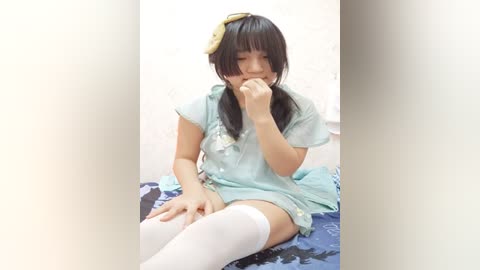 Media: Video of an East Asian woman with straight black hair, wearing a light blue dress, thigh-high white socks, and a yellow hair bow, sitting on a bed with blue and white sheets, covering her mouth with her hand.