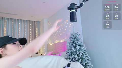 Video of a woman in a black cap, lying in a bed with a snowy tree and string lights, using a black microphone, in a cozy bedroom with pale walls.