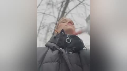 Media: A video of a young, fair-skinned woman with blonde hair, wearing a black puffy jacket, looking upward against a blurry, winter background with leafless trees.