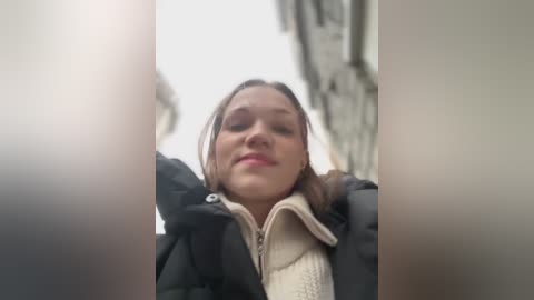 Media: A video captures a young woman with fair skin and brown hair, wearing a black jacket and beige scarf, looking upwards, with blurry, out-of-focus buildings in the background.