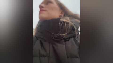 Media: A video captures a woman in a black puffy winter coat, with her head tilted back, possibly looking at something. The background is blurred, focusing on the coat's texture and her hair.