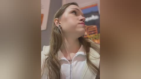 Media: Video of a young Caucasian woman with long brown hair, wearing a white shirt and beige jacket, gazing upward thoughtfully in an office setting with a blurred abstract painting in the background.