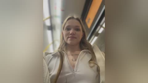 Media: Video of a young woman with long blonde hair, wearing a white jacket and a necklace, walking down a subway platform with blurred background.