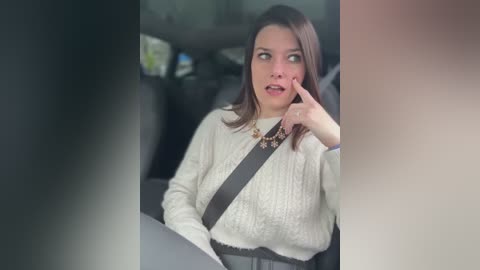 Media: Video of a Caucasian woman with shoulder-length brown hair, wearing a white knit sweater and black seatbelt, looking thoughtful with her finger on her lips, in a car.