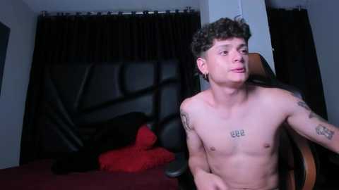 Media: Video of a shirtless young man with curly hair, tattoos, and piercings, standing in a dimly lit room with a black leather chair, a red pillow, and dark curtains in the background.