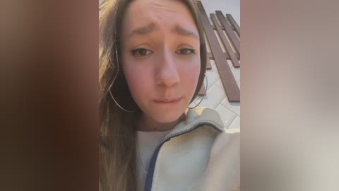 Media: A video of a young Caucasian woman with long brown hair, wearing a white jacket and hoop earrings, looking upset. Background shows a tiled floor with wooden planks.