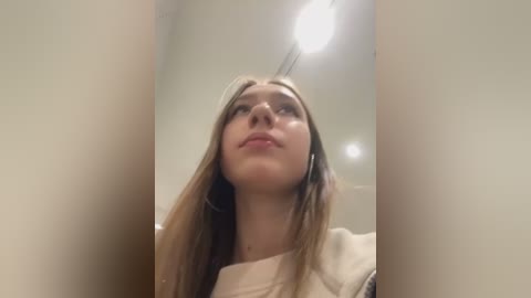 Media: A video of a young Caucasian woman with long, straight, light brown hair, wearing a beige sweater, looking upward with a neutral expression. The background is an indoor, well-lit space with a white ceiling.