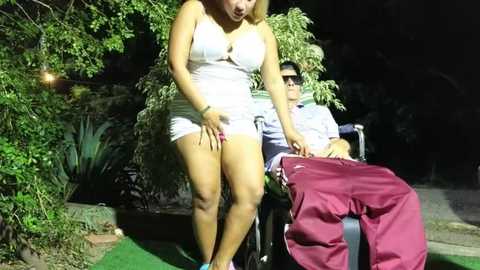 Media: Video of a curvy, light-skinned woman in a tight white dress, holding her crotch, standing next to a wheelchair-bound man in a dark shirt. Nighttime outdoor setting with lush greenery and a green carpet.