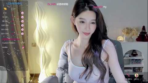 Media: Video of an East Asian woman with long dark hair, wearing a white tank top, sitting in a modern room with a white shelf and decorative items.