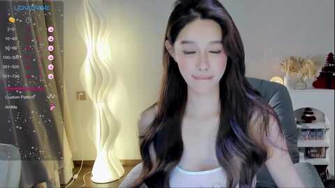 Media: A video of a young Asian woman with long dark hair, wearing a white tank top, seated in a dimly lit room with a modern lamp and a digital screen displaying social media icons.