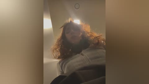 Media: A video of a young woman with curly red hair, wearing a light grey jacket, taking a selfie in a dimly lit elevator.