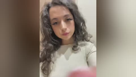 Video of a young woman with curly, dark hair and light skin, wearing a beige long-sleeved top. She has a neutral expression and is in a dimly lit room with blurred background.