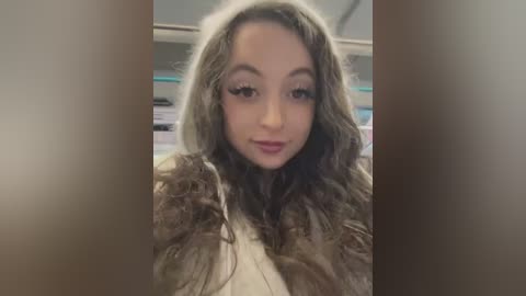 Media: A video of a young woman with wavy, shoulder-length brown hair, wearing a brown fur-trimmed jacket and a white top. She has a neutral expression and is indoors, with a blurred background of shelves and books.