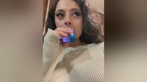 Video of a young woman with light skin, long dark hair, and full makeup, including dramatic eyeliner and mascara. She's wearing a cream-colored ribbed sweater, holding a blue and purple lipstick against her mouth. Background is blurred.